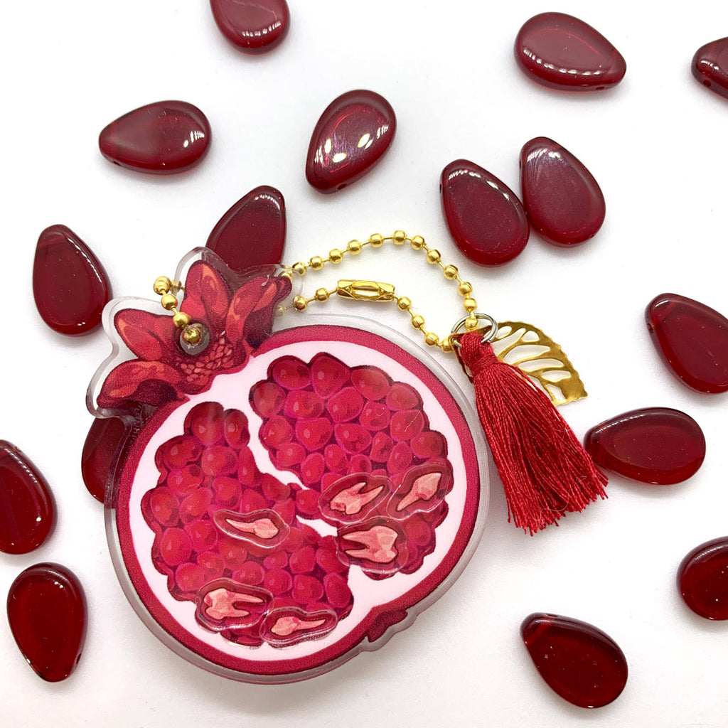 A transparent acrylic pomegranate charm with shakeable teeth inside, decorated with a gold beaded chain, a gold leaf, and a red tassel.
