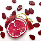 A transparent acrylic pomegranate charm with shakeable teeth inside, decorated with a gold beaded chain, a gold leaf, and a red tassel.