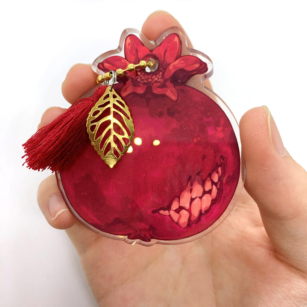 The backside of a transparent acrylic pomegranate charm with shakeable teeth inside, decorated with a gold beaded chain, a gold leaf, and a red tassel.